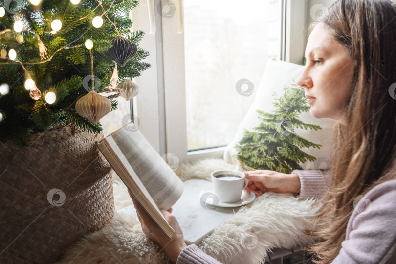 Скачать Женщина читает книгу и пьет кофе, сидя у окна в зимнем рождественском интерьере. фотосток Ozero