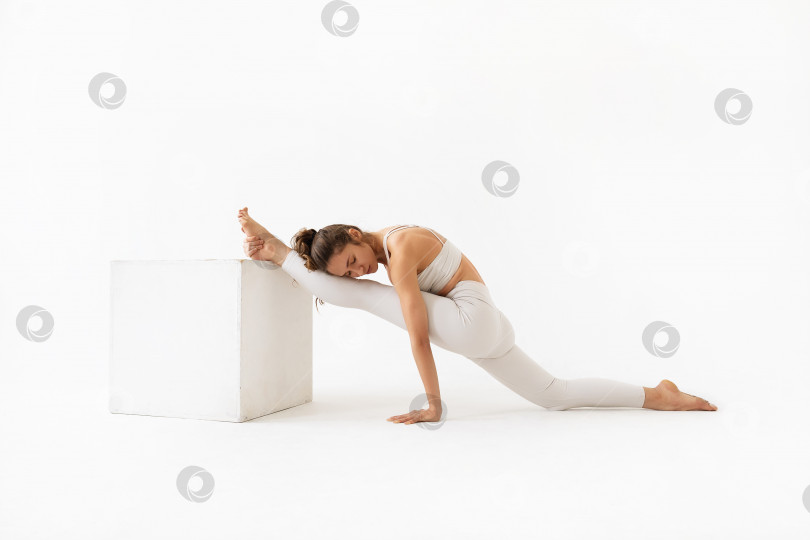Скачать Молодая женщина, занимающаяся йогой, выполняющая вариацию упражнения Хануманасана, продольный шпагат с опорой ноги на куб, тренируется в белой спортивной одежде в студии на светлом фоне. фотосток Ozero