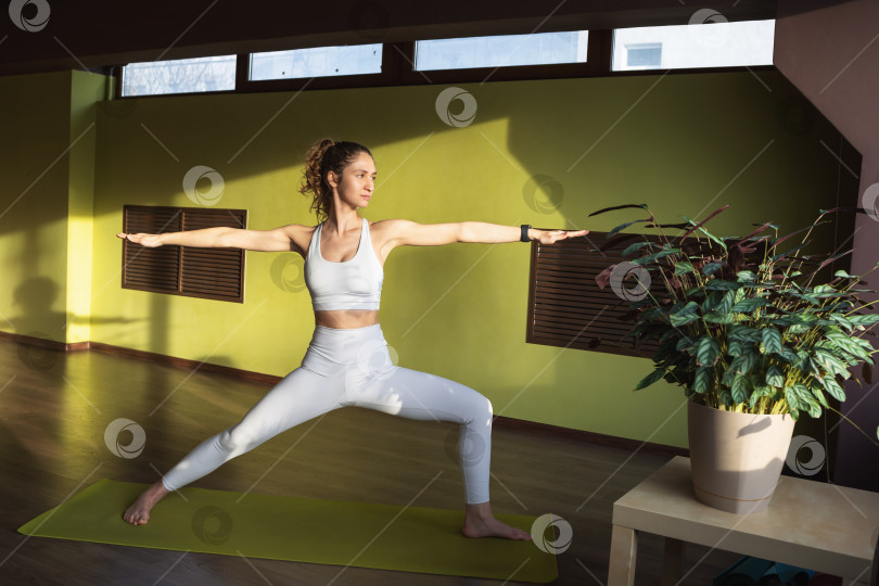 Скачать Молодая женщина, ведущая здоровый образ жизни и практикующая йогу, выполняет упражнение Вирабхадрасана, позу воина, тренируется в белой спортивной одежде в комнате на коврике фотосток Ozero
