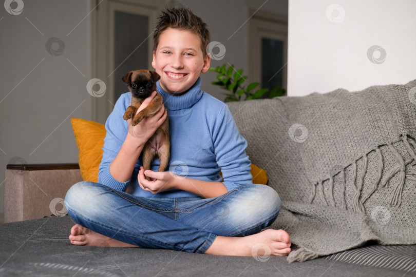 Скачать Мальчик играет с собачонкой. Малыш играет со щенком дома. Мальчик-подросток и собака породы гриффон или брабансон на диване. Дети и домашние животные дома. Ребенок сидит на полу с домашним животным. Уход за животными. фотосток Ozero
