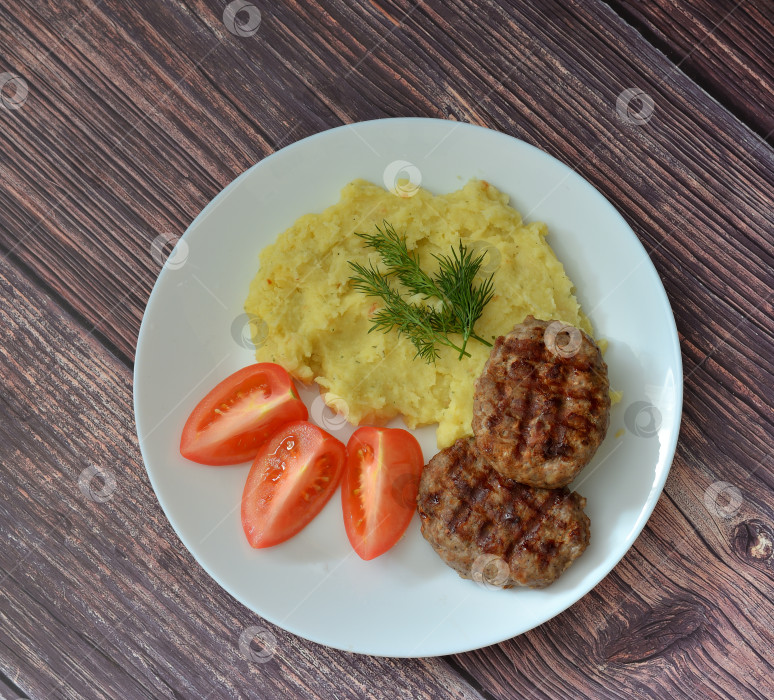 Скачать Две жареные мясные котлеты, картофельное пюре с зеленью и ломтиками помидоров на светлом деревянном столе. фотосток Ozero