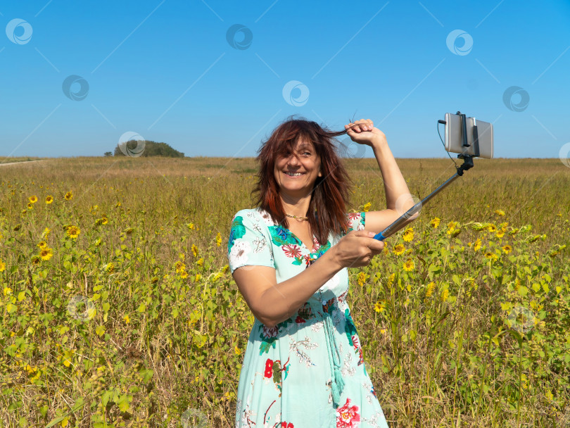 Скачать Улыбающаяся загорелая женщина в длинном платье на поле с подсолнухами делает селфи с помощью монопода фотосток Ozero