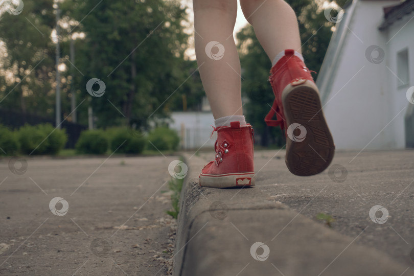 Скачать Девочка в красных кроссовках ходит по бордюру, развивает вестибулярный аппарат, равновесие. Прогулки и физические упражнения на свежем воздухе, теплым летним днем фотосток Ozero