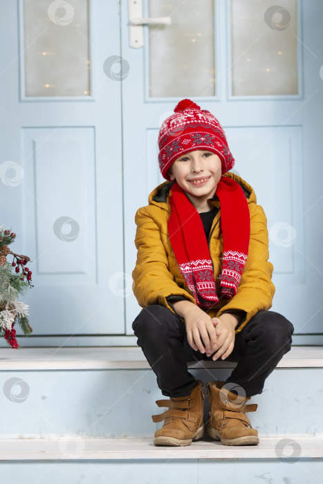 Скачать Красивый маленький мальчик в яркой зимней одежде сидит на крыльце дома. Счастливый ребенок зимой. фотосток Ozero