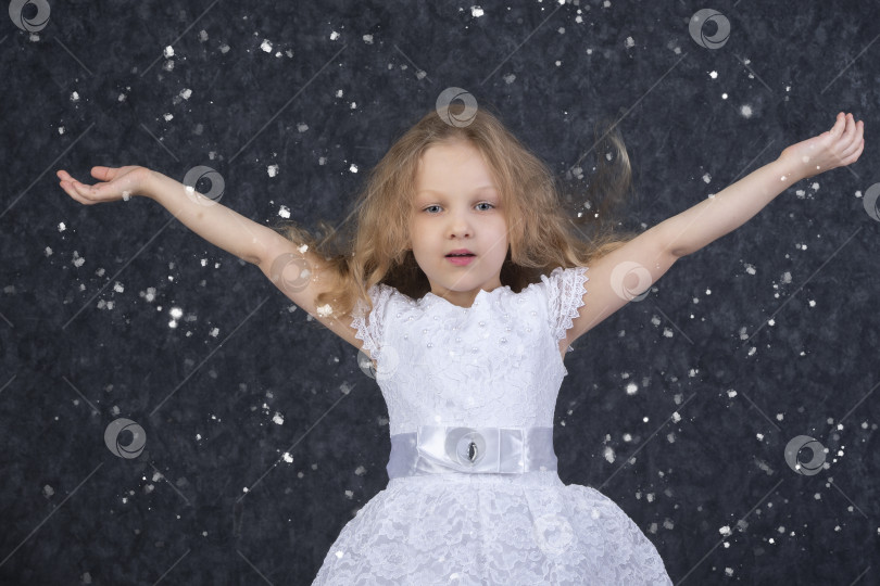 Скачать Красивая маленькая девочка в белом платье с поднятыми руками под летящими снежинками. фотосток Ozero