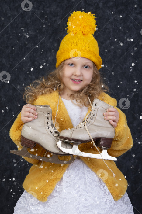 Скачать Счастливая маленькая девочка под белым снегом держит коньки в руках и улыбается. фотосток Ozero
