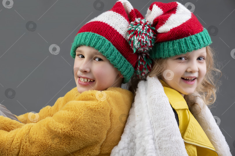 Скачать Красивые маленькие девочка и мальчик в вязаных рождественских шапочках сидят спиной друг к другу и улыбаются на сером фоне. Счастливого Рождества, дети. фотосток Ozero