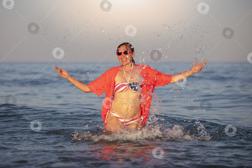 Скачать Счастливая женщина средних лет стоит в морской воде с поднятыми руками в купальнике с принтом американского флага. фотосток Ozero