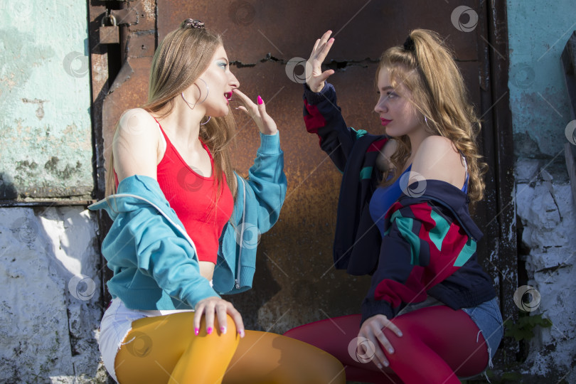 Скачать Две забавные девушки в яркой одежде в стиле девяностых сидят на фоне ржавой двери. Русские деревенские девушки. фотосток Ozero