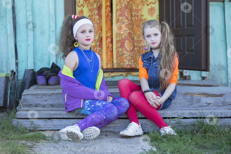 Скачать Две забавные маленькие девочки с ярким макияжем, одетые в стиле девяностых, сидят на деревенском крыльце дома. Русские деревенские дети. фотосток Ozero