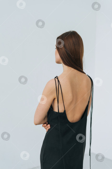 Скачать Модный портрет счастливой молодой женщины с длинными волосами в вечернем элегантном черном платье с открытой спиной в белом интерьере, поколение z, люди сзади фотосток Ozero