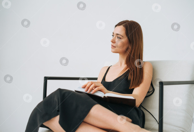 Скачать Красивая молодая женщина с темными длинными волосами в черном платье читает книгу на стуле в белом интерьере фотосток Ozero