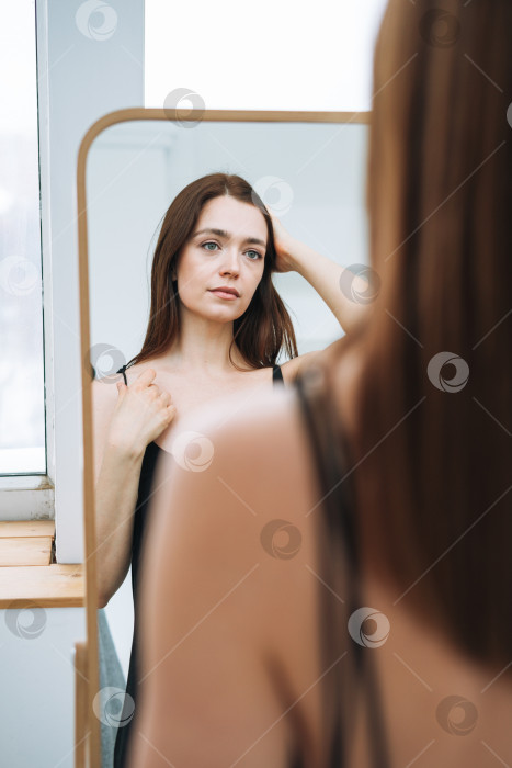 Скачать Молодая женщина с длинными темными волосами в вечернем элегантном черном платье с открытой спиной возле зеркала в белом интерьере фотосток Ozero