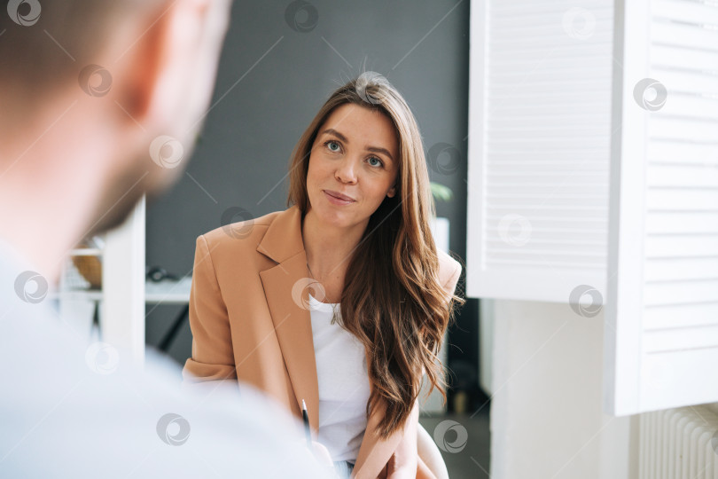 Скачать Молодая брюнетка с длинными волосами в стильном костюме работает с молодым человеком, женщина берет интервью у мужчины в светлом современном офисе фотосток Ozero