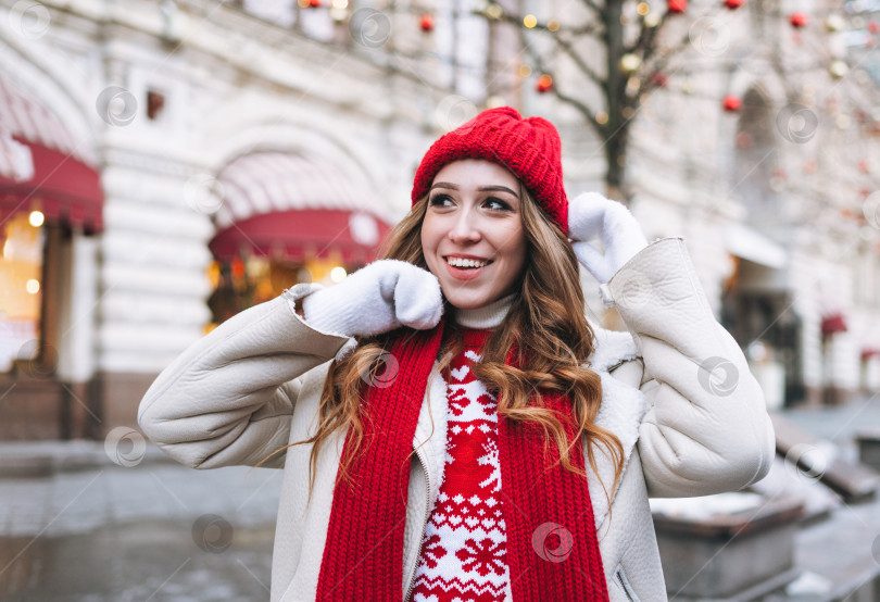 Скачать Молодая счастливая женщина с вьющимися волосами в красной вязаной шапочке на рождественской ярмарке на зимней улице, украшенной огнями фотосток Ozero