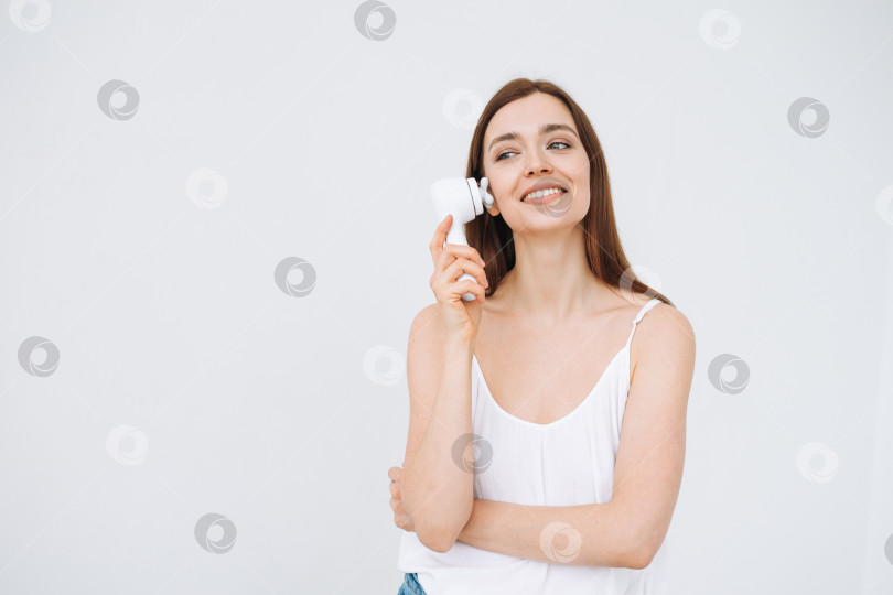 Скачать Красивый портрет счастливой улыбающейся женщины с темными длинными волосами с массажером для лица в руке на чистой свежей коже лица на белом фоне изолированно фотосток Ozero