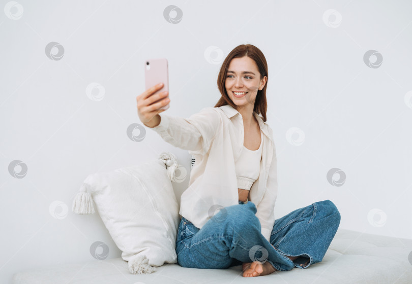 Скачать Красивая улыбающаяся женщина девочка подросток студентка с темными длинными волосами в белой рубашке делает селфи на мобильный телефон в руке, сидя на диване дома фотосток Ozero