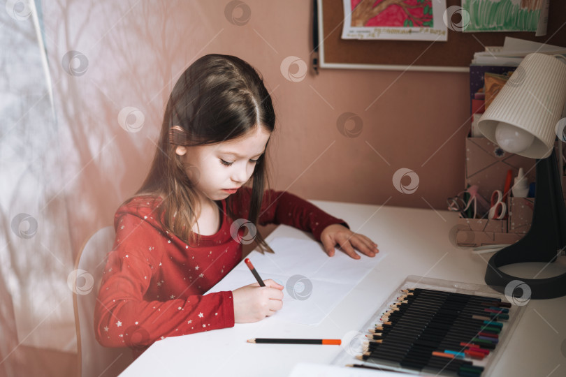 Скачать Симпатичная школьница с длинными волосами в красном платье рисует за столом в детской комнате дома фотосток Ozero