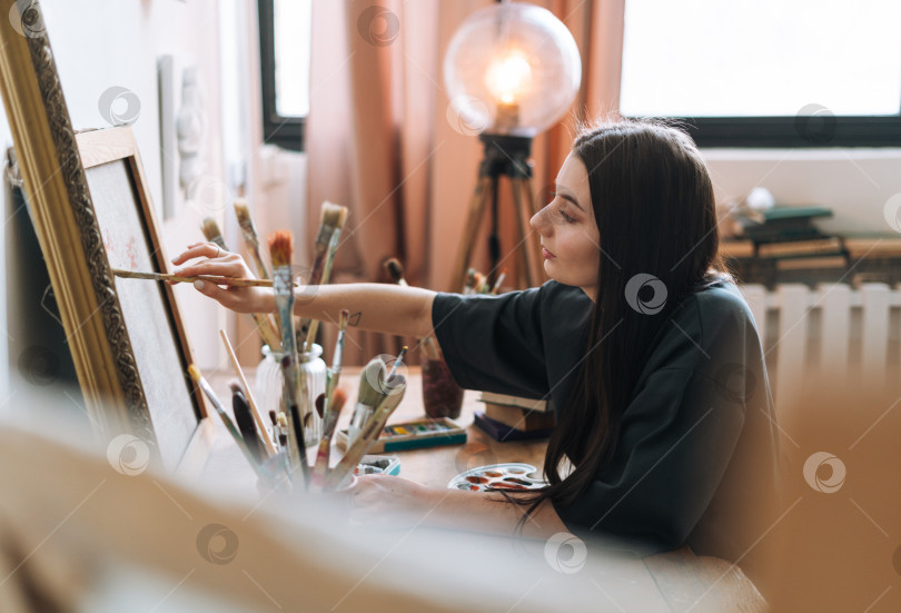 Скачать Молодая женщина-художник, девушка-подросток, студентка с темными длинными волосами в повседневной одежде рисует картину в художественной студии фотосток Ozero