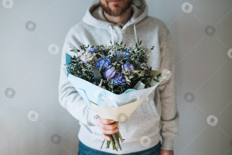 Скачать Молодой бородатый мужчина в серой толстовке с капюшоном и букетом цветов в руках на сером фоне фотосток Ozero