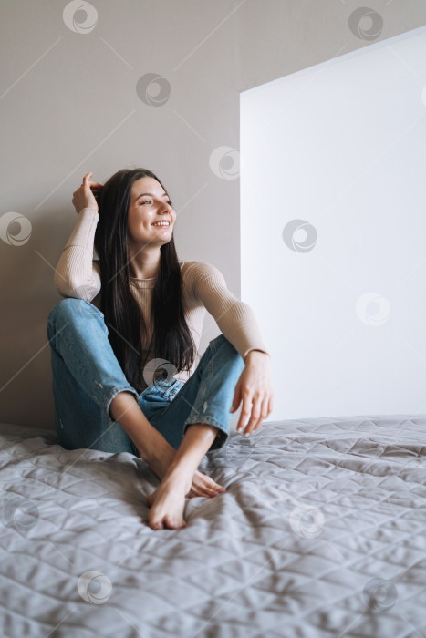 Скачать Молодая улыбающаяся женщина-подросток с темными длинными волосами в джинсах сидит на кровати в своей комнате фотосток Ozero