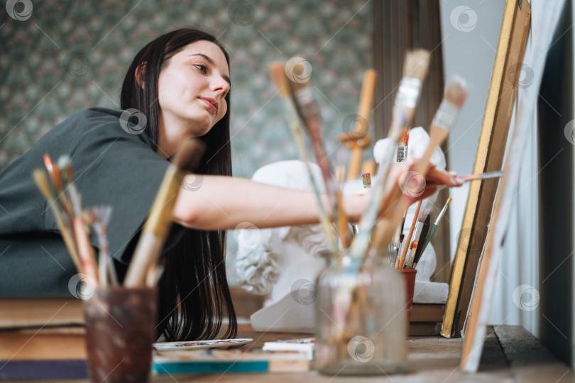 Скачать Молодая женщина-художник, девушка-подросток, студентка с темными длинными волосами в повседневной одежде рисует картину в художественной студии фотосток Ozero