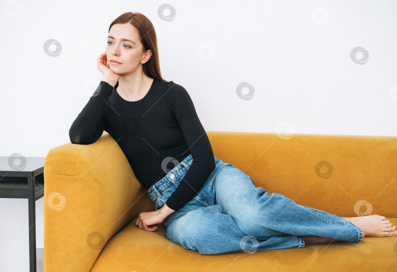 Скачать Молодая думающая женщина, девушка с темными длинными волосами в джинсах, сидит на желтом диване в минималистичном интерьере фотосток Ozero