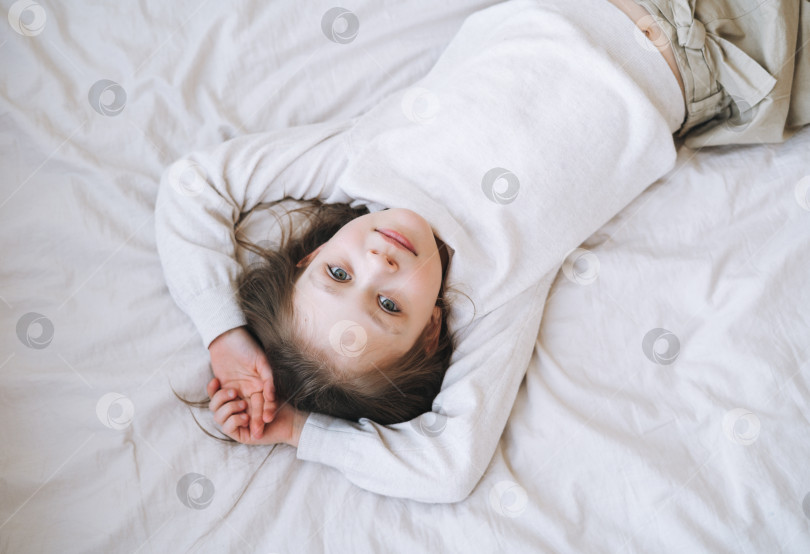 Скачать Симпатичная забавная маленькая девочка с длинными волосами в белом домашнем платье весело проводит время, лежа на кровати дома, вид сверху фотосток Ozero