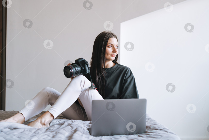 Скачать Молодая женщина-фотограф с цифровой камерой, фрилансер в повседневной одежде, использующая ноутбук, сидя дома на кровати фотосток Ozero