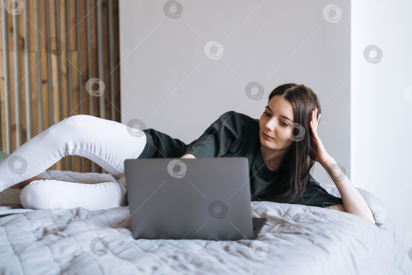 Скачать Красивая улыбающаяся женщина-подросток, студентка-фрилансер с темными длинными волосами в повседневной одежде, пользующаяся ноутбуком, сидя на кровати дома фотосток Ozero