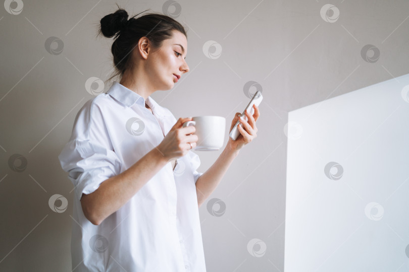 Скачать Красивая улыбающаяся женщина-подросток, студентка с темными длинными волосами в повседневной одежде, использующая мобильный телефон в руке дома фотосток Ozero