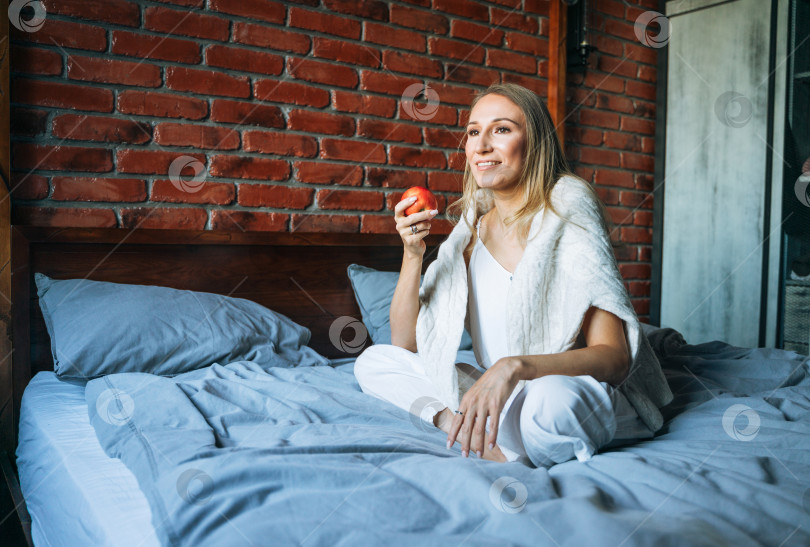 Скачать Молодая взрослая женщина со светлыми длинными волосами в повседневной одежде с яблоком сидит дома на кровати фотосток Ozero