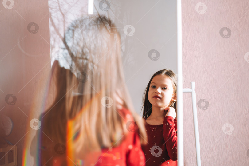 Скачать Симпатичная девочка-подросток с темными длинными волосами в красном платье смотрит в зеркало в детской комнате уютного дома фотосток Ozero