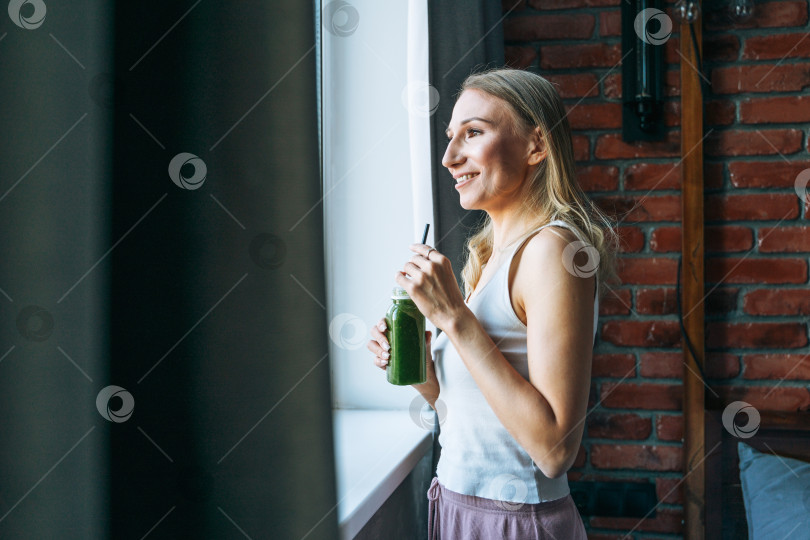 Скачать Молодая взрослая женщина со светлыми длинными волосами в повседневной одежде с зеленым коктейлем у окна дома фотосток Ozero