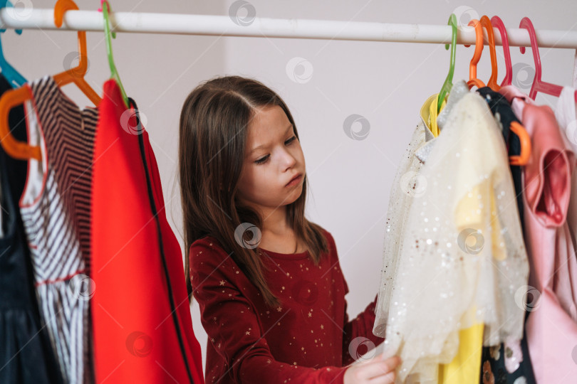 Скачать Размышляющая маленькая девочка с длинными темными волосами в красном платье среди своих красивых платьев в гардеробе в детской комнате дома фотосток Ozero