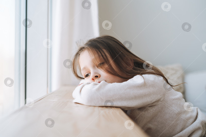 Скачать Симпатичная скучающе думающая маленькая девочка с длинными волосами в домашней одежде возле окна дома фотосток Ozero