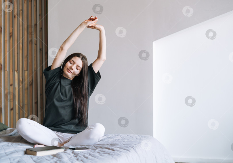 Скачать Молодая улыбающаяся женщина-подросток с темными длинными волосами в джинсах сидит на кровати в своей комнате фотосток Ozero