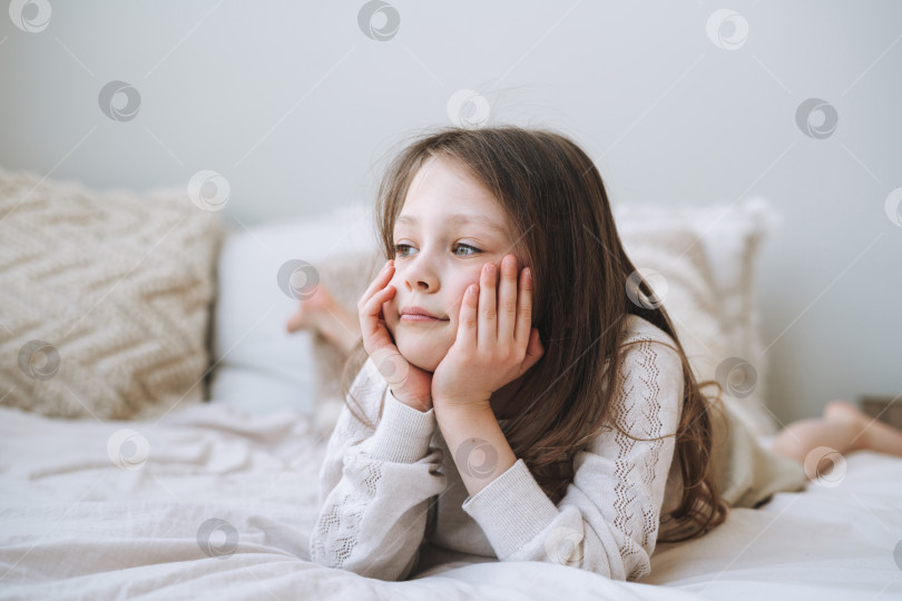 Скачать Симпатичная забавно мыслящая маленькая девочка с длинными волосами в домашней одежде весело проводит время, лежа на кровати дома фотосток Ozero
