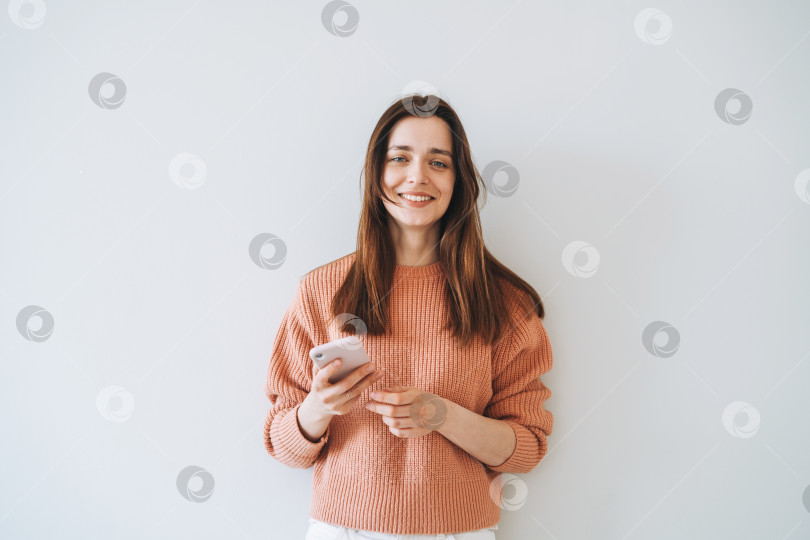 Скачать Портрет красивой улыбающейся студентки с темными длинными волосами в повседневном вязаном свитере с мобильным телефоном в руке на сером фоне фотосток Ozero