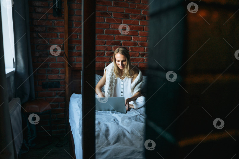 Скачать Взрослая женщина сорока лет-фрилансер со светлыми длинными волосами в повседневной одежде, пользующаяся ноутбуком, сидя на кровати дома фотосток Ozero