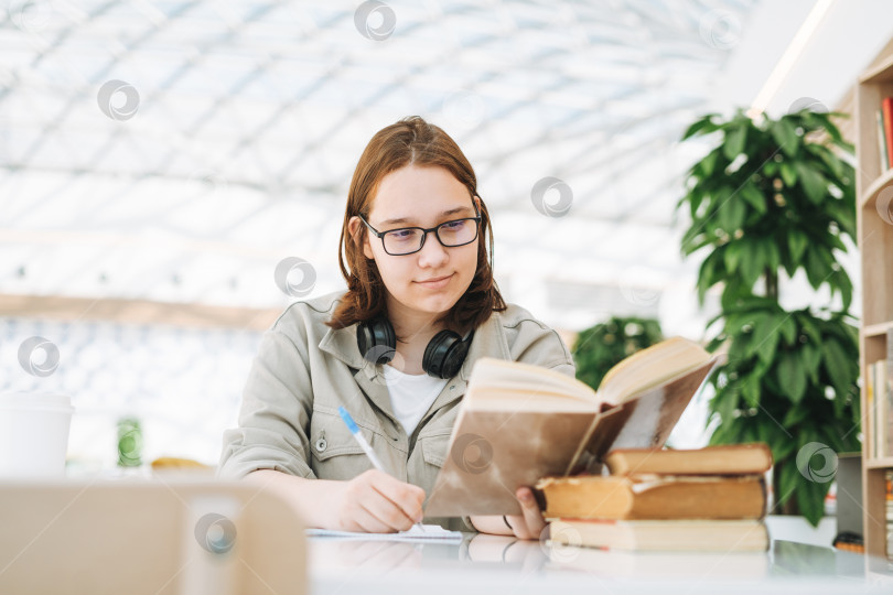 Скачать Молодая брюнетка-подросток, студентка колледжа в очках, делает домашнее задание с книгами в общественном месте green modern library фотосток Ozero