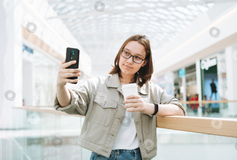 Скачать Молодая брюнетка студентка-подросток в очках с помощью мобильного телефона делает селфи с бумажным кофейным стаканчиком в общественном месте торгового центра фотосток Ozero