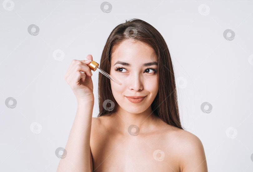 Скачать Красивый портрет счастливой улыбающейся азиатской женщины с темными длинными волосами с пипеткой с органическим маслом на чистой свежей коже лица и рук на белом фоне изолированно фотосток Ozero