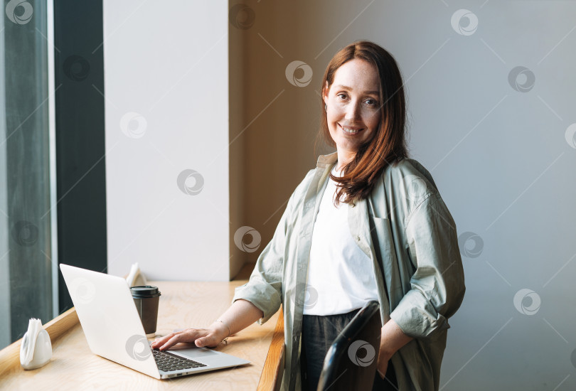 Скачать Взрослая улыбающаяся деловая женщина-брюнетка сорока лет с длинными волосами в стильной рубашке, работающая на ноутбуке в кафе фотосток Ozero