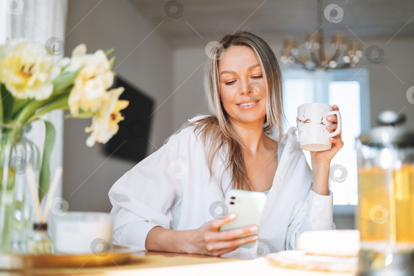 Скачать Молодая красивая счастливая женщина со светлыми длинными волосами в белой рубашке пьет чай с помощью мобильного телефона в солнечной гостиной дома фотосток Ozero