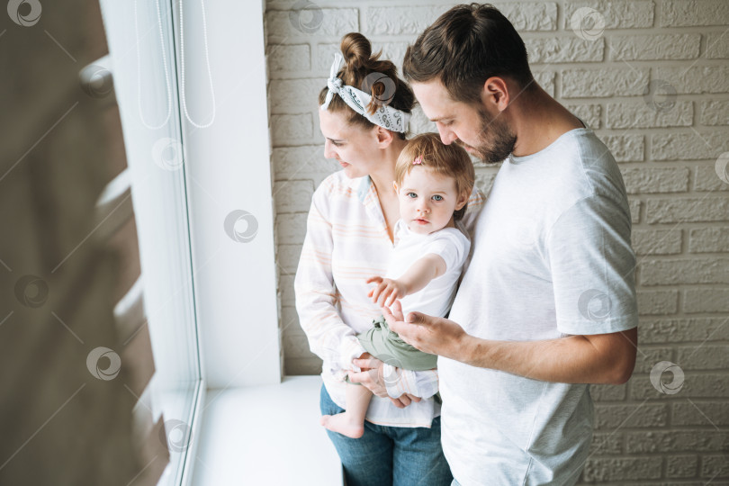 Скачать Молодой семьянин, отец и мать, женщина с маленькой девочкой на подоконнике, смотрят в окно дома фотосток Ozero
