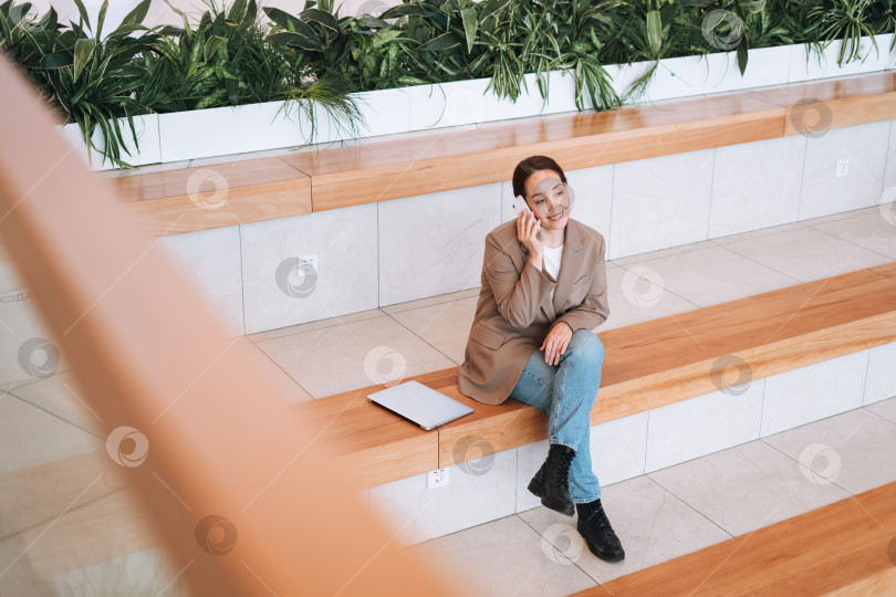 Скачать Взрослая улыбающаяся деловая женщина-брюнетка сорока лет с длинными волосами в стильном бежевом костюме и джинсах, работающая на ноутбуке в общественном месте фотосток Ozero