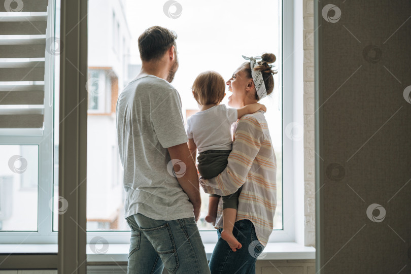Скачать Молодой семьянин, отец и мать, женщина с маленькой девочкой на подоконнике, смотрят в окно дома фотосток Ozero