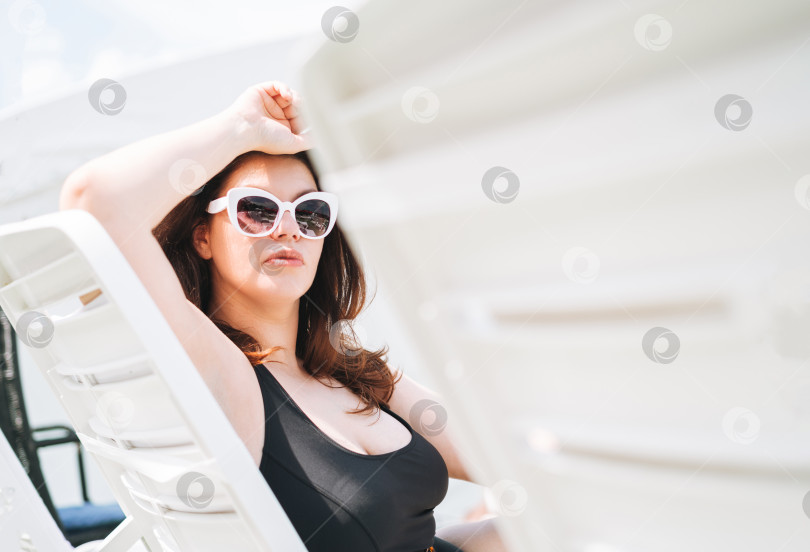 Скачать Стильная счастливая молодая женщина с позитивным телосложением плюс размер в черном купальнике и солнцезащитных очках, наслаждающаяся отдыхом на пляже в шезлонге у бассейна, летние каникулы фотосток Ozero