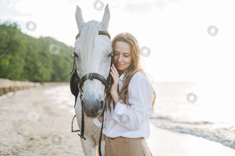 Скачать Молодая длинноволосая женщина в белой рубашке верхом на белой лошади на фоне морского пейзажа фотосток Ozero